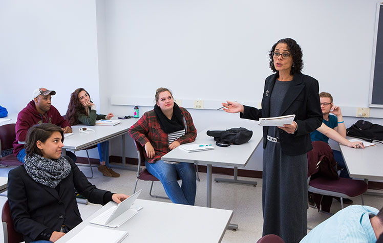 Classroom scene with professor talking while holding notebook and students looking on